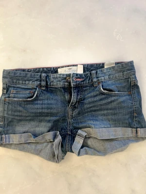 Blå jeansshorts med upprullad kant - Snygga blå jeansshorts med upprullad kant och klassisk femficksdesign. Shortsen har bälteshällor och knappgylf. Perfekta för varma dagar och ger en avslappnad look.