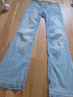Ljusblåa bootcut low waist jeans - Säljer ett par ljusblåa bootcut jeans med låg midja och slitna kanter nertill. Jeansen har en klassisk femficksmodell och är tillverkade i denim. Perfekta för dig som gillar retrovibbar och relaxed stil.                                    Midjemått: 32/33 cm.                                               Innerbenslängd: 81cm