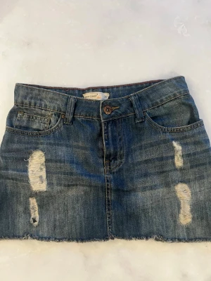 Kort jeanskjol med slitningar - Säljer en mörkblå kort jeanskjol i denim med slitna detaljer framtill och rå kant nertill. Kjolen har klassiska fem fickor, bälteshällor och knappstängning. Perfekt för dig som gillar en avslappnad och trendig stil.
