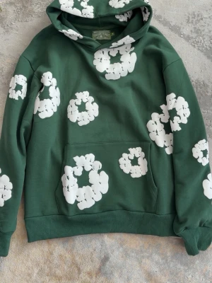 Grön hoodie från Denim Tears - Grön hoodie från Denim Tears med stora vita blommönster tryckta över hela plagget. Klassisk känguruficka framtill och ribbade muddar vid ärmar och nederkant. Tillverkad i mjukt bomullsmaterial och har en rymlig huva. Sitter lite större