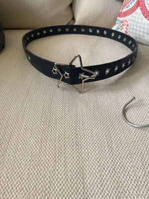Svart bälte med stjärnspänne - Snyggt svart bälte med silverfärgade nitar och ett unikt stjärnformat spänne i metall. Bältet har små stjärndetaljer längs hela remmen och passar perfekt för att ge din outfit en cool touch.