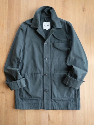 COS Dark Green Chore Jacket, size S/M - Stilren mörkgrön overshirt från COS i kraftig bomull. Jackan har tre utanpåliggande fickor, knäppning framtill och klassisk krage. Perfekt lager-på-lager-plagg med avslappnad passform och uppvikta ärmar för en cool look.
