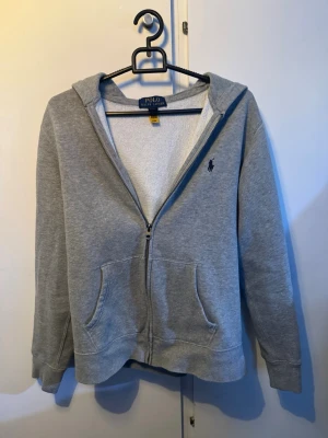 Grå zip hoodie från Polo Ralph Lauren - Snygg grå hoodie med dragkedja från Polo Ralph Lauren. I typ helt ny skicke har inga märken eller skavanker. Passar de typ 165 skulle jag säga. Om priset inte duget kom med bud.