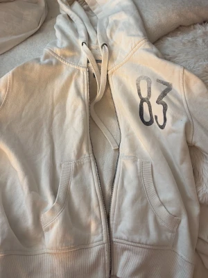 Vit sip hoodie  - Vit sip hoodie, i ett urtvättat tryck '83' i svart på bröstet, två framfickor och ribbade ärmmuddar och nedtill. 
