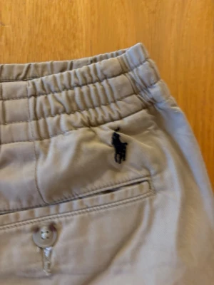 Beige chinos med resår från Polo Ralph Lauren - Snygga beige chinos från Polo Ralph Lauren med resår i midjan och dragsko. Klassisk rak passform och diskreta bakfickor med knapp. Tillverkade i mjukt bomullstyg och dekorerade med den ikoniska loggan broderad ovanför fickan.