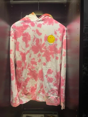 Rosa tie-dye hoodie med smiley - Säljer en hoodie i rosa och vit tie-dye med stor gul smiley och texten 'Positive State of Mind' på ryggen. Mindre smiley på bröstet. Hoodien har huva och magficka, samt ribbade muddar. Perfekt för dig som gillar färg och positiv vibe.