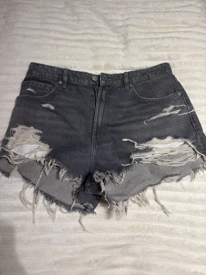 Gråa slitna high waist shorts - Säljer ett par gråa högmidjade jeansshorts med rejält slitna detaljer och fransig kant. Shortsen har klassisk femficksmodell och knappgylf. Perfekta för dig som gillar en edgy och avslappnad stil.
