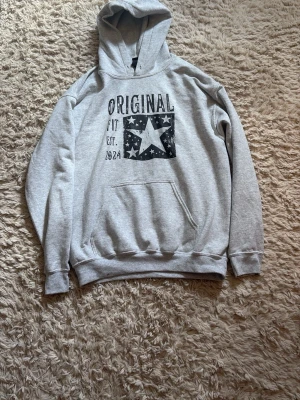 Grå hoodie med tryck och huva - Säljer en grå hoodie med svart tryck framtill där det står 'ORIGINAL' och en stor stjärna, från original archive Hoodien har huva och en stor magficka. Mjuk och skön, perfekt till chill dagar. Klassisk passform och ribbade muddar vid ärmar och nederkant. Knappt använd pga den var för stor när jag köpte den.