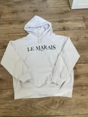 Vit hoodie med LE MARAIS-tryck - Säljer en vit hoodie med svart text 'LE MARAIS' på bröstet. Hoodien har huva med dragsko, magficka och ribbade muddar vid ärmar och nederkant. Perfekt för en avslappnad och trendig look. Materialet känns mjukt och bekvämt. Storlek 140 