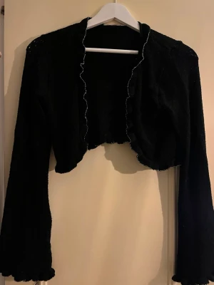Svart stickad bolero med volangkant - Snygg svart stickad bolero med volangkant längs öppningen och vid ärmsluten. Kort modell med långa ärmar, perfekt att slänga över en topp eller klänning för en extra touch. Enkel och stilren design som passar till mycket.