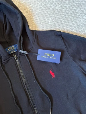Svart hoodie från Polo Ralph Lauren - Snygg svart hoodie med dragkedja från Polo Ralph Lauren. Klassisk design med huva, fickor framtill och det ikoniska röda Polo-logot broderat på bröstet. Tillverkad i mjukt material som känns skönt mot huden. Perfekt för en avslappnad och stilren look.