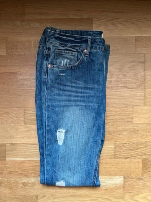 Blå bootcut jeans  - Säljer ett par blå jeans med bootcut passform och slitningar på låret och vid benslut. Jeansen har klassisk femficksdesign och är gjorda i denim med en snygg tvättad look. Perfekta för en avslappnad och trendig stil.