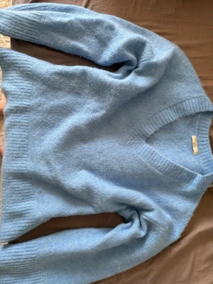 Ljusblå stickad tröja Gina Tricot - Mysig ljusblå stickad tröja från Gina Tricot med v-ringning och ribbade muddar vid ärmslut och nederkant. Tröjan har en fluffig och mjuk känsla, perfekt för kyliga dagar. Enkel och stilren design som är lätt att matcha.