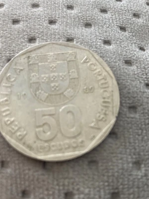 Portugisisk 50 escudos mynt - Säljer ett portugisiskt 50 escudos mynt i silverfärg med landets vapensköld på ena sidan och ett skepp på andra sidan. Myntet är från 1989 och har en kantig form. Perfekt för samlare eller dig som gillar unika utländska mynt.