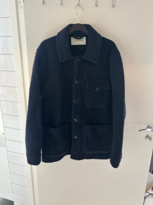 Mörkblå ull overshirt med fickor - Stilren mörkblå overshirt i ullmaterial med klassisk krage och knäppning framtill. Jackan har tre stora fickor, varav en bröstficka med lock och två nedtill. Perfekt lager på lager-plagg för kyligare dagar. Storlek S men passar M