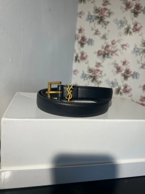 Svart bälte med guldspänne YSL - Snyggt svart bälte i skinn med guldfärgat YSL-spänne. Perfekt accessoar för att lyfta vilken outfit som helst. Klassisk design med exklusiv känsla och stilren finish.