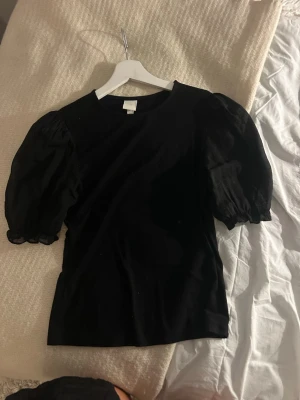 Svart topp med puffärmar - Snygg svart topp med puffiga kortärmar och rund halsringning. Toppens material känns mjukt och stretchigt, perfekt för en trendig look. Passformen är normal och toppen är enkel att matcha med olika stilar. 
