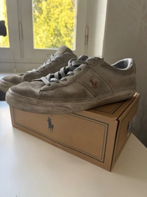 Gråa sneakers från Polo Ralph Lauren - Snygga gråa sneakers i mocka från Polo Ralph Lauren med klassisk låg profil. Skorna har grå snörning, diskret logga på sidan och POLO-text bak på hälen. Perfekta för en clean och stilren look. Har använts, därför priset är lågt. ABS! Pris går att diskuteras vid snabb affär!