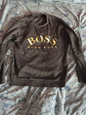 Hugo boss tröja - Svart sweatshirt från Hugo Boss med stor guldfärgad logga på bröstet. Tröjan har rund halsringning, långa ärmar och ribbade muddar vid ärmslut och nederkant. Tillverkad i mjukt bomullsmaterial för en bekväm känsla.