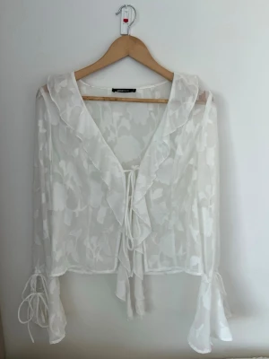 Vit transparent blus med volang - Snygg vit transparent blus med blommigt mönster, volangkantad krage och knytband framtill. Långa ärmar med utsvängda ärmslut och dekorativa knytband vid handlederna. Perfekt för en trendig och luftig look.