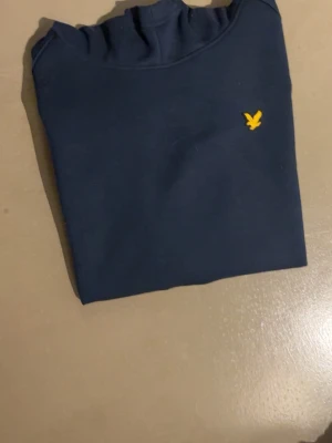 Lyle scott hoodie - Snygg mörkblå hoodie med en liten gul broderad logga på bröstet. Klassisk modell med huva och långa ärmar. Perfekt för en chill och avslappnad stil. Materialet känns mjukt och bekvämt mot huden.