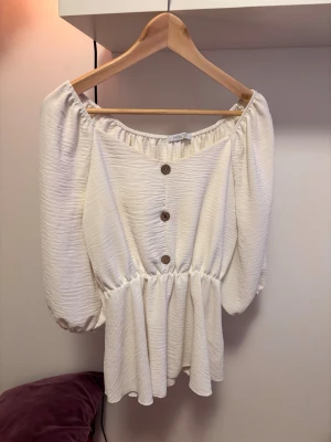 Shein Offshoulder cremefärgad blus med knappar - Trendig cremefärgad offshoulder-blus med puffiga långärmar och träknappar framtill. Blusen har en markerad midja och ett flowy fall nedtill. Materialet är lätt strukturerat och ger en cool, avslappnad vibe.
