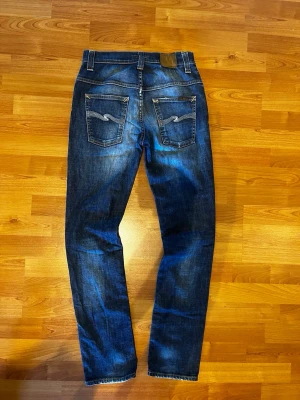 Mörkblå nudie slim jeans  - Säljer ett par mörkblå slim jeans med slitningar på både fram- och baksida. Jeansen har klassiska fem fickor och kontrastsömmar. Materialet är denim i bomull och passformen är lite avslappnad men ändå snyggt åtsittande.