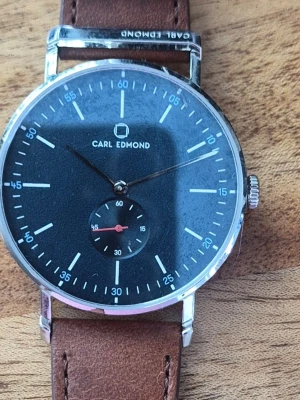 Carl Edmond klocka med brun läderrem - Stilren klocka från Carl Edmond med svart urtavla, silverfärgat boett och bruna läderband. Urtavlan har minimalistiska vita markeringar och en liten sekundvisare. Perfekt accessoar för dig som gillar klassisk och modern design.