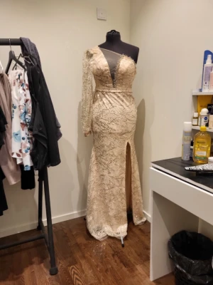 Champagnefärgad långklänning med slits - Elegant långklänning i champagnefärg med glittriga paljettdetaljer och pärlor. Klänningen har en unik asymmetrisk ärm och en hög slits framtill. Perfekt för dig som vill sticka ut med en lyxig och glamorös look. Mått: byst ca 90cm, midja ca 77cm höft ca 103cm längd 150cm från axel. Tyget är ej strechigt.