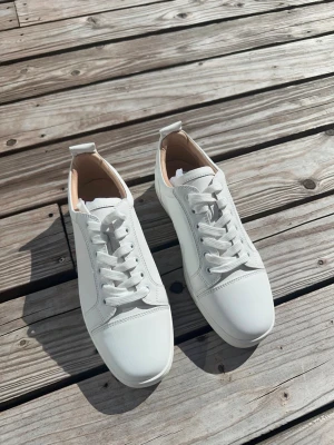 Vita Louboutin  - Stilrena vita sneakers i läder från Christian Louboutin med klassisk låg profil och ikonisk röd sula med logotyp. Skorna har vita snören och diskreta sömmar för en clean look. Perfekta för dig som vill ha något exklusivt och tidlöst.