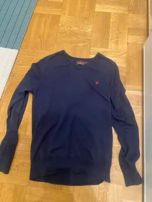 Mörkblå V-ringad tröja - Mörkblå V-ringad tröja med lång ärm och ribbade muddar. Liten röd broderad logga på bröstet ger en snygg detalj. Perfekt till jeans eller chinos för en clean look.