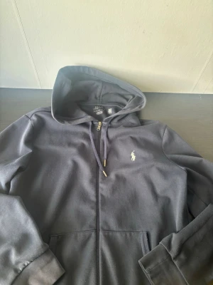 Mörkblå hoodie från Polo Ralph Lauren - Snygg mörkblå hoodie från Polo Ralph Lauren med dragkedja framtill och klassisk broderad logga på bröstet. Hoodien har fickor på sidorna, justerbar huva med snören och ribbade muddar vid ärmar och nederkant. Tillverkad i mjukt bomullsmaterial.