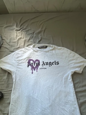 Vit Palm Angels t-shirt med tryck - Säljer en vit t-shirt från Palm Angels med svart text och ett lila hjärta med drip-effekt på bröstet. Modellen är klassisk med rund hals och korta ärmar. Perfekt för dig som gillar streetwear och vill sticka ut lite extra.