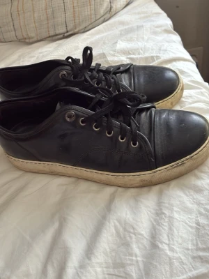 Svarta låga sneakers med vita sulor - Snygga svarta sneakers med lågt skaft och vita, slitstarka gummisulor. Skorna har svarta skosnören och metallfärgade öljetter. Ovandelen är i slätt material som ger en clean look, perfekt till jeans eller streetwear.