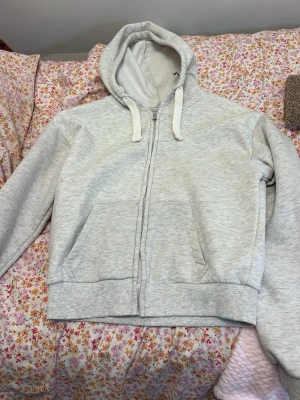 Gina tricot kofta - En basic ljusgrå hoodie med dragkedja framtill och två stora fickor. Hoodien har justerbar huva med snören och ribbade muddar vid ärmar och nederkant. Perfekt för chill dagar eller när du vill ha en enkel och clean look.