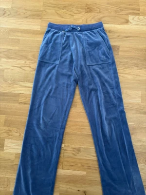 JUICY COUTURE Blå mjukisbyxor i velour - Säljer ett par blå mjukisbyxor i mjuk velour med bred resårmidja och snörning. Byxorna har raka ben och stora fickor framtill. Perfekta för chill dagar eller när du vill vara extra bekväm.