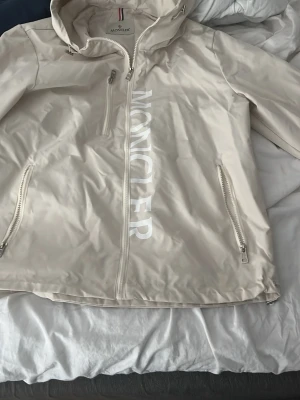 Beige vindjacka från Moncler - Snygg beige vindjacka från Moncler med stor vit logga längs dragkedjan. Jackan har huva, tre dragkedjeförsedda fickor och är tillverkad i ett glansigt, lätt material som passar perfekt för blåsiga dagar. Stilsäker och enkel att matcha till olika outfits.är orginelt storlek l men passar som M