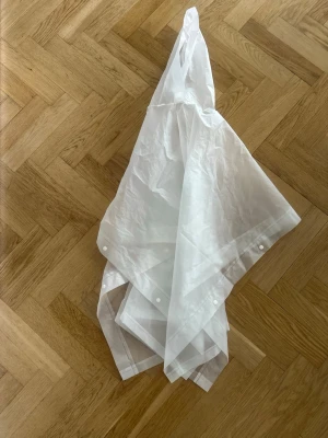 Vit regnponcho Kolmården - Genomskinlig vit regnponcho med tryck från Kolmården. Ponchon har huva och knappar på sidorna för enkel på- och avtagning. Tillverkad i lätt plastmaterial, perfekt att slänga på sig när det börjar regna.