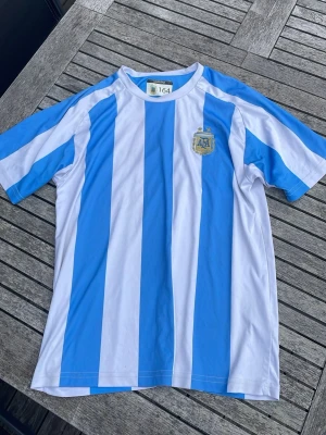 Argentina fotbollströja blåvit - Säljer en klassisk Argentina fotbollströja med breda blå och vita vertikala ränder. Tröjan har korta ärmar och rund hals, samt Argentinas fotbollsförbunds märke på bröstet. Perfekt för dig som vill stötta landslaget på match eller träning.