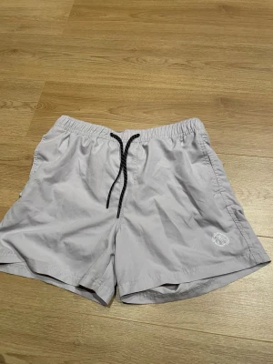 Ljusgrå badbyxor från Jack & Jones - Säljer ett par ljusgrå badbyxor från Jack & Jones med svart dragsko i midjan och elastisk resår. Byxorna har en liten vit logga på vänster ben och meshfoder på insidan. Perfekta för stranden eller poolen.