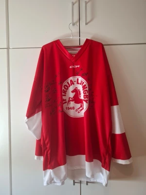 Röd IF Troja-Ljungby hockeytröja + merch - Säljer en röd och vit hockeytröja från IF Troja-Ljungby med klubbens logga och årtal 1948 på bröstet. Tröjan har V-ringning, långa ärmar och är signerad av flera spelare. Tillverkad i ett slitstarkt syntetmaterial, perfekt för hockeyfans.