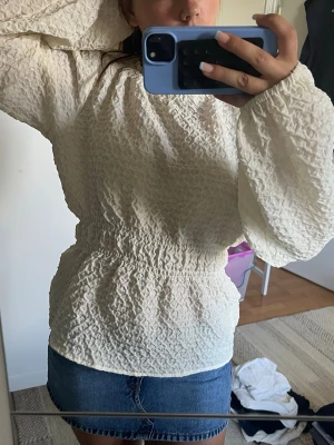 Krämvit blus med puffärmar - Supersnygg krämvit blus med långärmade puffärmar och smockad midja. Blusen har en strukturerad, skrynklig yta som ger en cool och trendig look. Perfekt att styla med jeans eller kjol för en chic outfit.