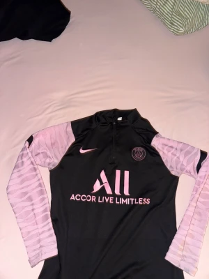 Svart och rosa PSG träningströja Nike - Snygg långärmad träningströja från PSG i svart med rosa detaljer och mönstrade ärmar. Halvlång dragkedja framtill och klubbmärke på bröstet. Tillverkad i ett lätt och funktionellt material som passar perfekt till träning.
