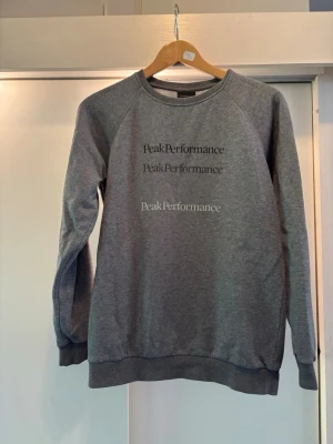 Grå sweatshirt Peak Performance - Snygg grå sweatshirt från Peak Performance med tryckt logga i tre olika nyanser på bröstet. Tröjan har rund halsringning, långa ärmar och ribbade muddar vid ärmslut och nederkant. Perfekt för en avslappnad och sportig stil🏋️🍋‍🟩