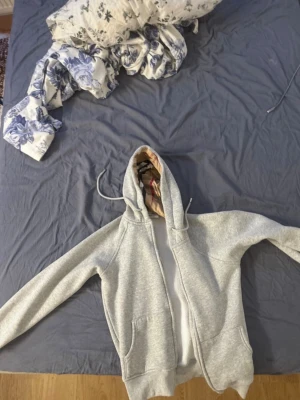 Grå hoodie med dragkedja och mönstrad huva - Säljer en grå hoodie med dragkedja och fickor framtill. Insidan av huvan har ett snyggt rutigt mönster i beige, svart och rött. Perfekt för en chill och avslappnad stil. Dragsko i huvan för extra passform.