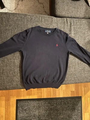 Mörkblå stickad tröja Polo Ralph Lauren - Mörkblå stickad tröja från Polo Ralph Lauren med rund halsringning och klassisk röd broderad logga på bröstet. Tröjan har långa ärmar och ribbade muddar vid ärmslut och nederkant. Perfekt för en stilren och tidlös look.