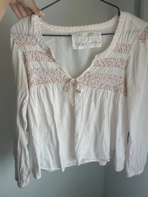 Vit blus med broderi från Odd Molly - Söt vit blus från Odd Molly med ljusrosa broderade detaljer och smock upptill. Blusen har v-ringning med liten rosett och långa ärmar. Tillverkad i ett tunt, luftigt material som känns skönt mot huden. Perfekt för en avslappnad men ändå stilsäker look.