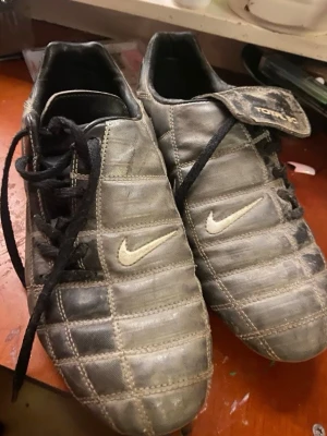 Grå Nike fotbollsskor med dobbar - Säljer ett par klassiska grå fotbollsskor från Nike med vita detaljer och snörning. Skorna har tålig ovandel i syntetmaterial och dobbar för bra grepp på planen. Perfekta för dig som vill ha stabilitet och stil på matchen. Behöver tvättas 