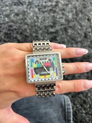 Färgglad fyrkantig klocka från D&G - Unik fyrkantig klocka från D&G med metallarmband och färgstarkt testbildsmönster på urtavlan. Klockan har en cool retrostil med silverfärgad boett och detaljer i flera färger som blå, gul, röd och grön. Perfekt för dig som vill sticka ut! 34mm tror jag. Behöver nytt batteri 