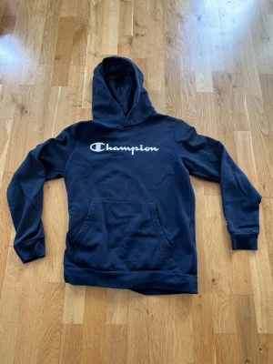  Champion hoodie med logga - 162cl. Snygg mörkblå hoodie från Champion med stor vit logga över bröstet. Klassisk känguruficka framtill och huva. Perfekt för en avslappnad streetstyle-look. Mjuk bomullskänsla och ribbade muddar vid ärmslut och nederkant.